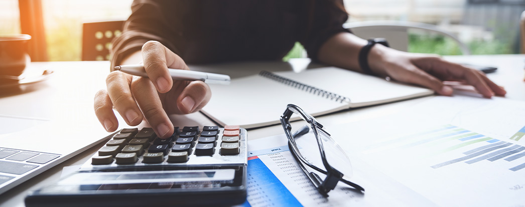 mãos segurando caneta e digitando na calculadora, papeis a mesa, caderno e óculos